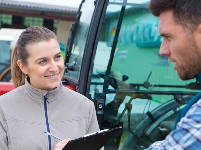 A woman standing smiling and holding clipboard as a male farmer talks to her.